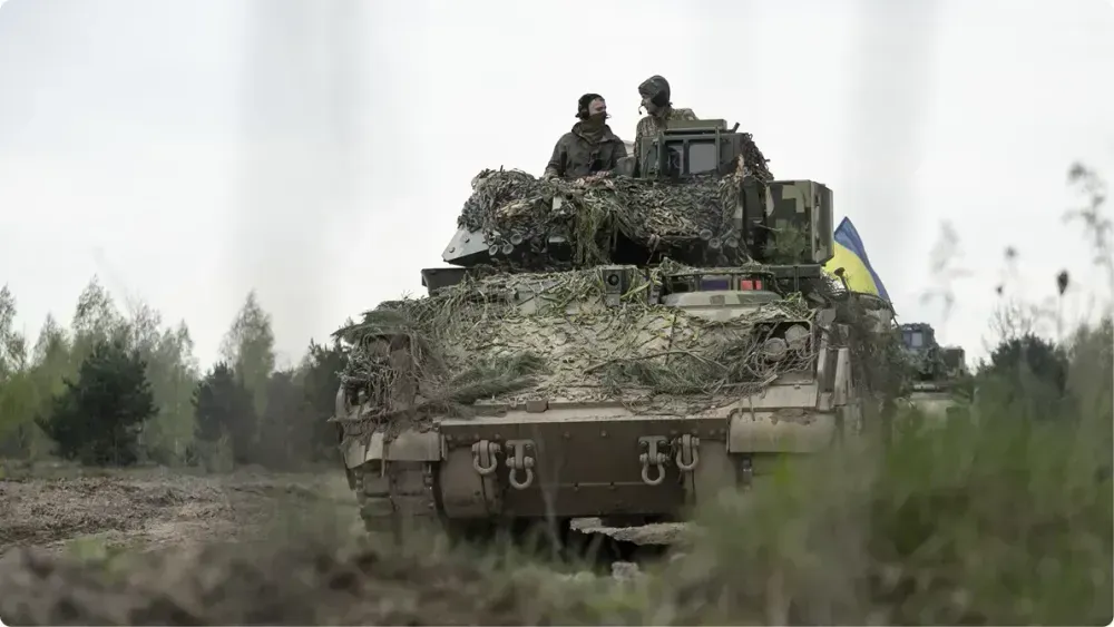 Два українські військовослужбовці стоять у відкритому люку основного бойового танка, вкритого маскувальною сіткою та гілками, під час руху груntовою дорогою. На борту техніки видно жовто-синій прапор України. На задньому плані — ще одна одиниця техніки.